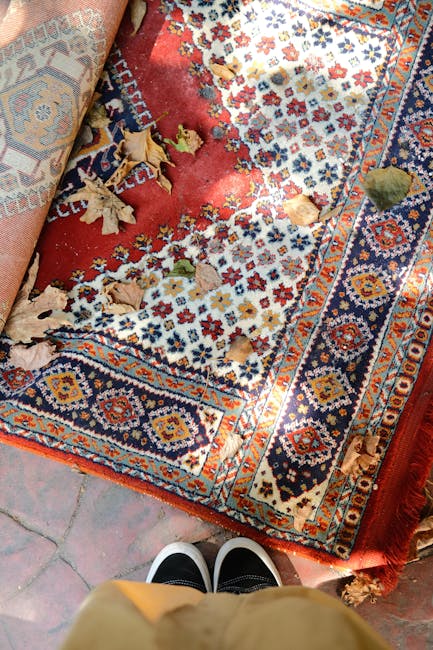 A broom with a straw bristle head rests against a patterned rug, which is draped over a plain white wall in a corner of a room. The rug features geometric and traditional motifs, partially covering a