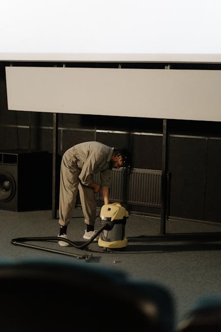 A yellow canister vacuum cleaner with a long black hose and attachment is placed on a patterned area rug in a living room with wooden flooring. In the background, there is a black media console with a