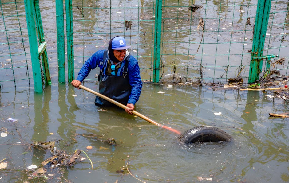 A worker in waterproof clothing, including a blue jacket and a blue baseball cap, is standing knee-deep in murky floodwater behind a green metal fence. The worker is using a long-handled tool, likely 