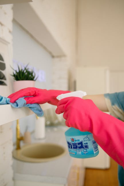 A person cleaning a smooth, dark tiled floor in a bright, modern living room. The individual is dressed in a gray sweater, black pants, and white socks, operating a flat mop with a gray handle and a w